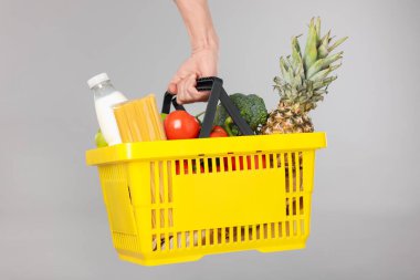 Elinde gri arka planda farklı ürünler olan alışveriş sepeti tutan adam, yakın plan.