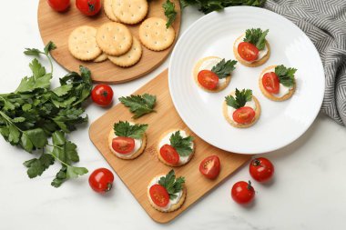 Krem peynirli krakerler, domatesler ve maydanozlar beyaz masada.