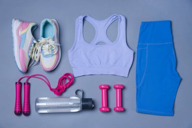 Spor kıyafetleri, spor ayakkabıları, ip atlama, dambıl ve su şişesi gri arka planda, düz yatış.