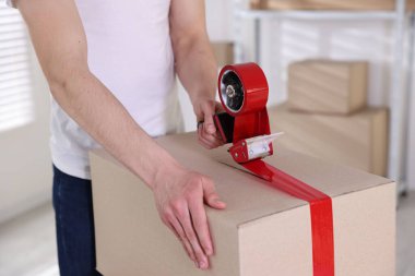 Karton kutuyu kapalı bir yere kapatmak için yapıştırıcılı bant kullanan bir adam.