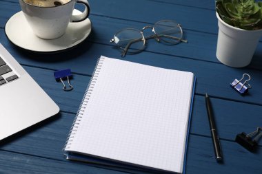 Kalemli, dizüstü bilgisayarlı, gözlüklü, ataçlı ve mavi ahşap masadaki kahveli defter.
