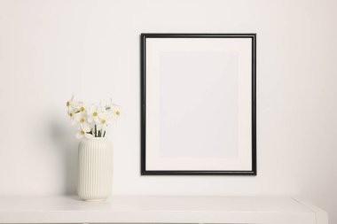 Beyaz masadaki vazodaki güzel nergisler ve kapalı alandaki resim çerçeveleri