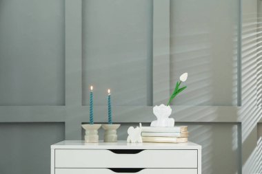 Gri duvarın yanındaki çekmecelerin üzerinde yanan mumlar olan şamdanlar.