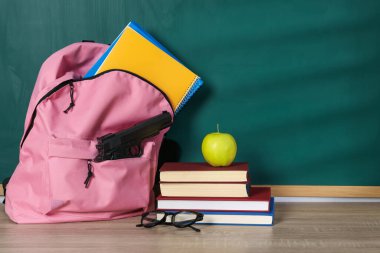 Sırt çantasında silah, kırtasiye malzemeleri, bardaklar ve elma tahtanın yanındaki tahta masada.