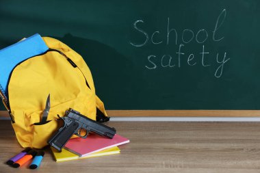 Sırt çantasında silah, bıçak ve kırtasiye malzemeleri var. Tahtanın yanındaki tahta masada. Okul güvenliği