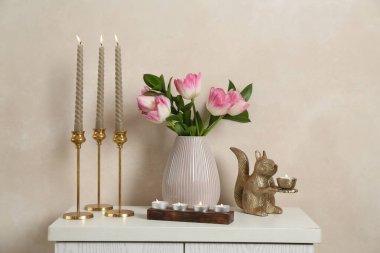 Mumları yanan zarif şamdanlar vazoda laleler ve bej duvarın yanındaki şifoniyerde diğer dekorlar.