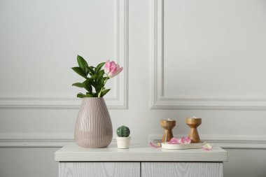 Zarif şamdanlar, yanan mumlar, vazoda lale ve beyaz duvarın yanındaki dolapta diğer dekorlar.