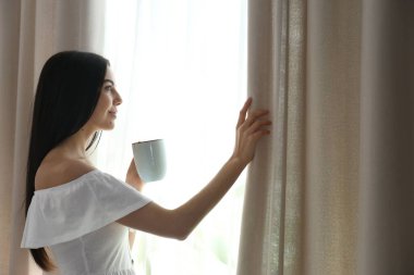 Woman with cup of coffee opening window curtain at home in morning
