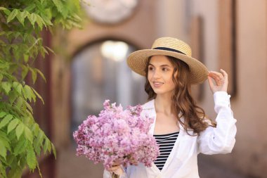 Şapkalı gülümseyen kadın, şehir caddesindeki ağacın yanında bir demet leylak çiçeğiyle.