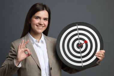 Hedef alınıyor. Dart tahtası ve okla gülümseyen iş kadını gri arka planda iyi bir hareket gösteriyor.