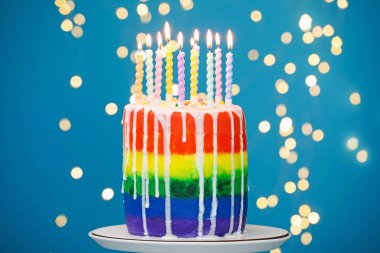 Enfes gökkuşağı doğum günü pastası açık mavi arka planda yanan mumlar, bulanık ışıklar, yakın plan