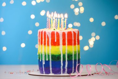 Enfes gökkuşağı doğum günü pastası yanan mumlar, serpiştirmeler ve flama ile mavi arka planda bulanık ışıklar, yakın plan