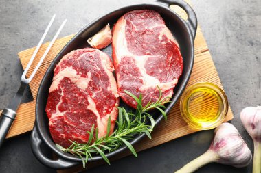 Fırın kabında baharatlı çiğ et parçaları, gri desenli masa üzerinde yağ ve çatal, düz yatar.