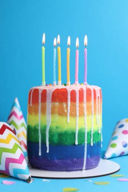 Enfes gökkuşağı doğum günü pastası yanan mumlar, parti şapkaları ve açık mavi arka planda konfeti