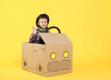 Küçük çocuk sarı arka planda kartondan yapılmış arabayı kullanırken el sallıyor. Metin için boşluk
