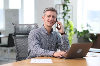 Gülümseyen işadamı akıllı telefondan konuşuyor ve ofiste dizüstü bilgisayarla çalışıyor.