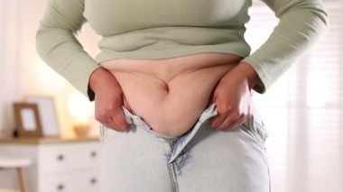 Kilolu bir kadın evde dar kot pantolon giymeye çalışıyor.