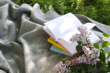 Yığınla kitap, leylak çiçekleri ve yeşil çimlerin üzerinde battaniye.