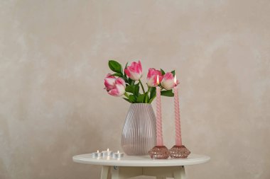 Bej duvarın yanındaki beyaz masada yanan mumlar ve vazolarda laleler olan zarif tutucular.