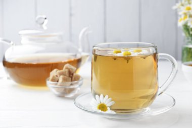 Cam fincanda nefis papatya çayı, kahverengi şeker, çaydanlık ve beyaz ahşap masada taze çiçekler, yakın plan.