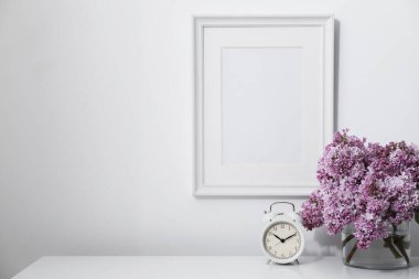 Beyaz duvarın yanındaki masada güzel leylak çiçekleri ve çalar saat var.