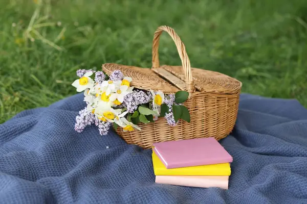 Yığınla kitap, battaniye ve hasır sepet. Yeşil çimlerin üzerinde çiçekler.