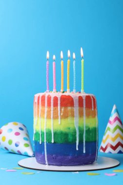 Enfes gökkuşağı doğum günü pastası yanan mumlar, parti şapkaları ve açık mavi arka planda konfeti