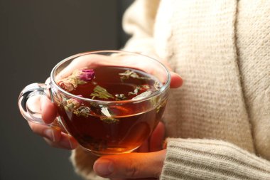 Gri arka planda cam bardakta aromatik bitki çayı olan kadın, yakın plan.