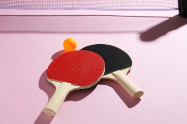 Masa tenisi raketleri, top ve net pembe arka planda, görünümün üstünde.