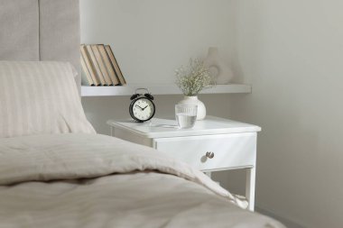 Komodinin yanında çalar saat, vazoda çiçekler ve yatağın yanında bir bardak su.