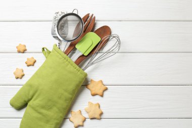 Fırın eldiveninde farklı mutfak aletleri ve beyaz ahşap masada yıldız şeklinde kurabiyeler. Metin için boşluk