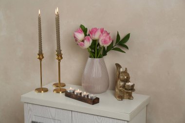 Mumları yanan zarif şamdanlar vazoda laleler ve bej duvarın yanındaki şifoniyerde diğer dekorlar.