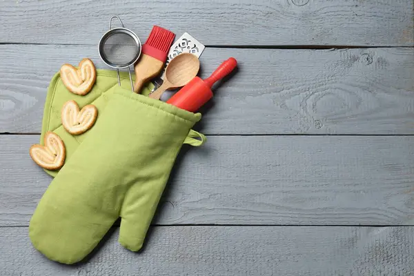 Fırın eldiveninde farklı mutfak gereçleri ve açık gri ahşap masada düz bir şekilde duran avuç içi kurabiyeler. Metin için boşluk