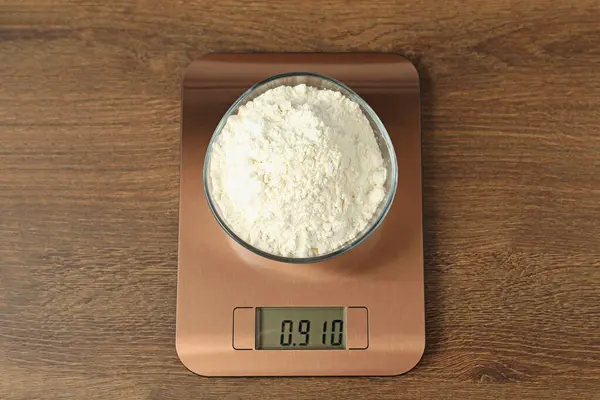 Modern electronic kitchen scale with bowl of flour on wooden table, top view