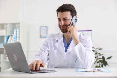 Hastanedeki beyaz masada dizüstü bilgisayarla çalışırken akıllı telefondan konuşan profesyonel bir doktor.