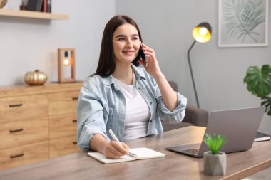 Planlayıcılı gülümseyen kadın notlar alıyor ve ahşap masada akıllı telefondan konuşuyor.