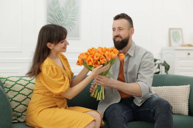 Bir adam evdeki kanepede sevgili karısına güzel laleler sunuyor.