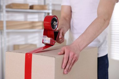 Karton kutuyu kapalı bir yere kapatmak için yapıştırıcılı bant kullanan bir adam.