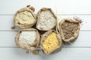 Beyaz ahşap masa üzerinde kağıt torbalarda farklı türde un, düz yerde.