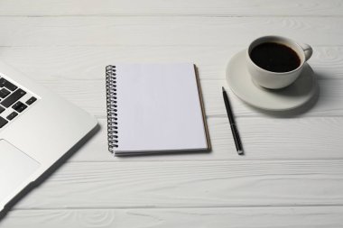 Beyaz ahşap masada defter, kalem, kahve ve dizüstü bilgisayar.