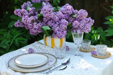 Bahçedeki masada güzel leylak çiçekleri, tabaklar, çatal bıçaklar, bardaklar ve mumlar
