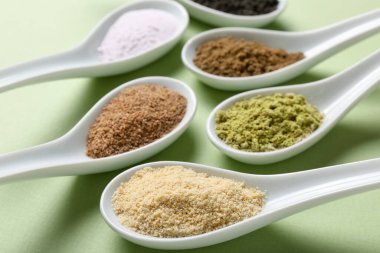 Açık yeşil arka planda kaşıklarda farklı süper yiyecek tozları, yakın plan.