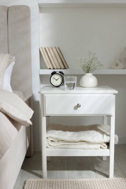 Komodinin yanında çalar saat, vazoda çiçekler ve yatağın yanında bir bardak su.