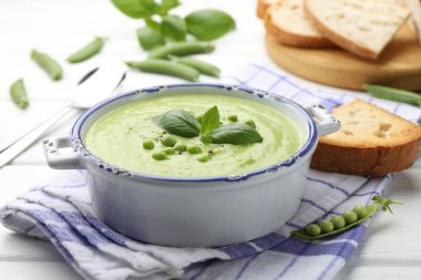 Beyaz ahşap masada servis edilen fesleğenli bezelye kremalı çorba.