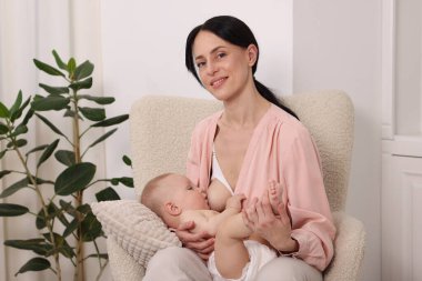 Gülümseyen anne evdeki koltukta bebeğini emziriyor.