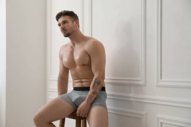 Beyaz duvarın yanındaki sandalyede şık, açık gri iç çamaşırlı genç adam. Metin için boşluk
