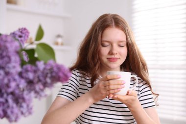 Evinde bir fincan sıcak içeceği ve leylak çiçekleri olan güzel bir genç kız.