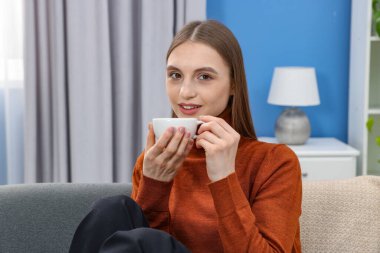 Evdeki kanepede bir fincan kahveyle gülümseyen kadın.