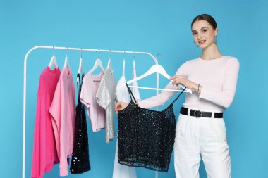 Gülümseyen moda tasarımcısı ve açık mavi arka planda giysileri olan askılar.