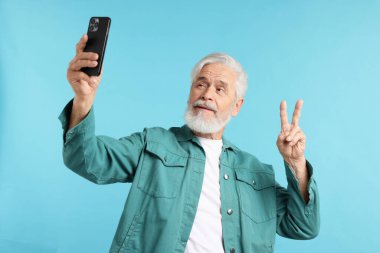 Açık mavi arkaplanda selfie çekerken huzur işareti gösteren yaşlı bir adam